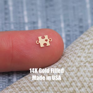 May include: A gold-filled puzzle piece charm with a heart cutout. The charm is shown on a finger and the text "14K Gold Filled Made in USA" is visible below.