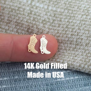 May include: Two small gold-colored cowboy boot charms. One charm is gold-filled, and the other is silver-colored. The charms are detailed with decorative stitching. The text "14K Gold Filled Made in USA" is visible.