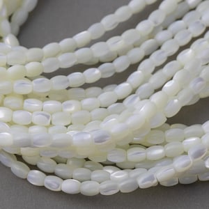 Op de afbeelding: Een streng witte ovale parelmoer kralen. De kralen hebben een licht iriserende glans.