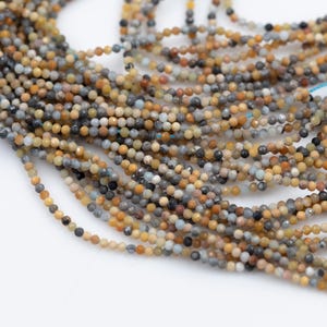 May include: Strands of small, round beads in various colors, including tan, gray, and black, are displayed against a white background. The beads appear to be strung together, creating a textured and detailed surface. The image suggests a craft or jewelry-making theme.