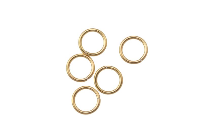 May include: Five gold-colored jump rings are scattered against a white background. Each ring is circular with a small opening, suggesting they are designed for jewelry making or crafts. The rings have a smooth, shiny finish.