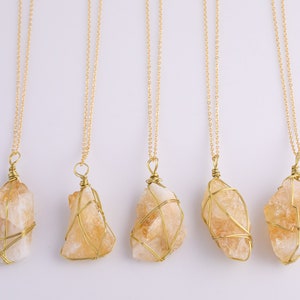 May include: Five citrine crystal pendants on gold chains. Each raw citrine stone is wrapped in gold wire. The necklaces are displayed against a white background. The citrine stones are a light orange colour.