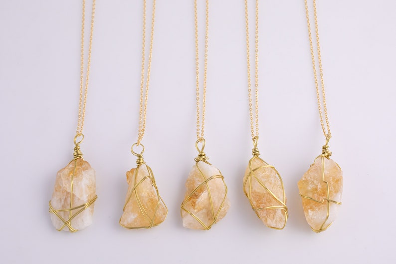 May include: Five citrine crystal pendants on gold chains. Each raw citrine stone is wrapped in gold wire. The necklaces are displayed against a white background. The citrine stones are a light orange colour.