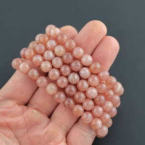 May include: A strand of pink sunstone beads, each bead is round and smooth. The beads are strung on a clear elastic cord.