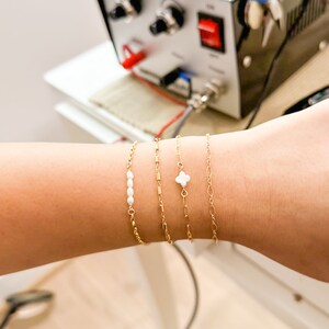 May include: A close-up of a wrist wearing four gold bracelets. One bracelet has a small white clover charm, another has a row of small white pearls, and the other two are simple gold chains.