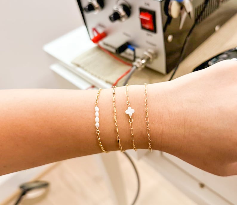 May include: A close-up of a wrist wearing four gold bracelets. One bracelet has a small white clover charm, another has a row of small white pearls, and the other two are simple gold chains.