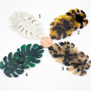 May include: Four pairs of plastic monstera leaf shapes in different colors and patterns. The leaves are white with a marbled pattern, brown with a tortoise shell pattern, brown with a mottled pattern, and green with a marbled pattern.