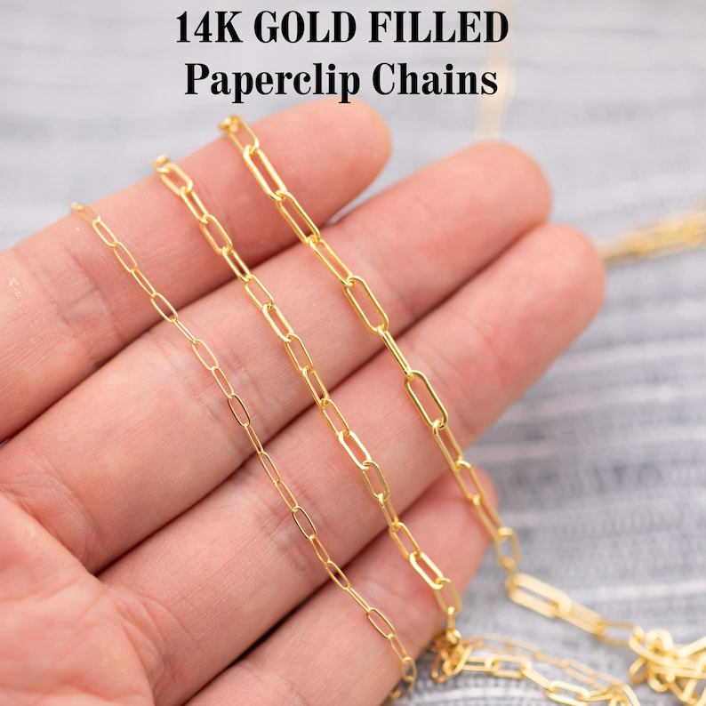 May include: Close-up of three gold-filled paperclip chains. The chains are displayed against a neutral background. The text "14K GOLD FILLED Paperclip Chains" is at the top of the image. The chains vary in size and link shape.