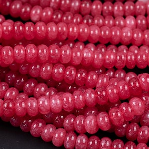 May include: Close-up of strands of red-pink gemstone beads. The beads are small, rounded, and have a polished, glossy finish. The strands are arranged in a layered pattern, creating a textured surface. The color is a vibrant, saturated red-pink.