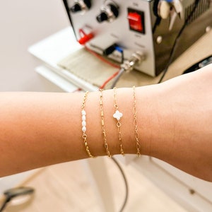 May include: A close-up of a wrist wearing four gold bracelets. One bracelet has a small white clover charm, another has a row of small white pearls, and the other two are simple gold chains.