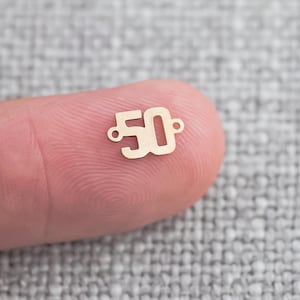 May include: A gold-colored metal charm shaped like the number 50. The charm has two small holes for attaching it to a necklace or bracelet.