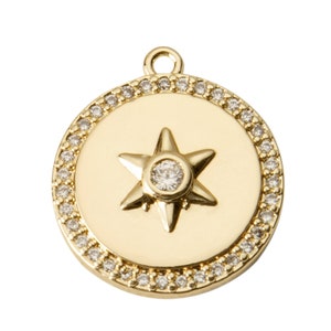 May include: A gold-colored pendant with a star design. The pendant features a central star with a clear gemstone and is surrounded by a ring of smaller, clear gemstones. The pendant has a loop at the top for attaching to a chain.