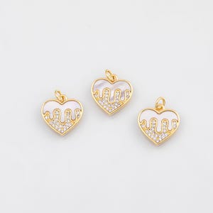 Puede incluir: Tres colgantes en forma de corazón con marcos dorados. Cada colgante presenta un centro blanco nacarado con un diseño dorado que gotea y una fila de cristales transparentes. Los colgantes tienen pequeños bucles en la parte superior para colgar.