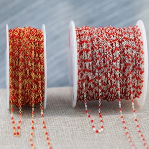 May include: Two spools of beaded chain. One spool features gold chain with red beads, the other has silver chain with red beads. The chains are wound around white spools, with strands of the chain hanging down.
