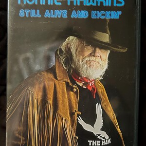 May include: A DVD case featuring a portrait of Ronnie Hawkins, a country music singer. The case has a black background with blue text that reads "Ronnie Hawkins Still Alive and Kickin'" and a white bird logo with the text "The Hawk".