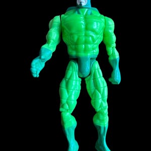 May include: A green action figure with a muscular physique and a blue head covering. The figure has a stern expression and is posed against a black background. The figure's arms and legs are articulated.