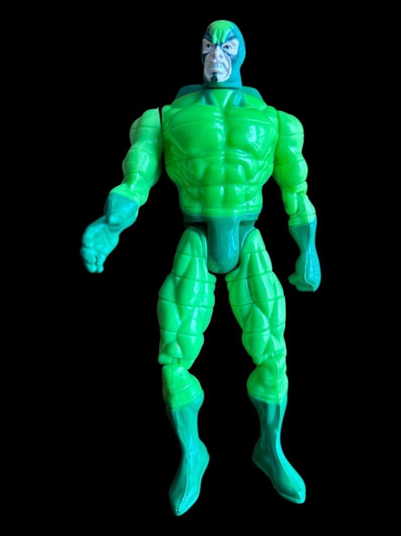 May include: A green action figure with a muscular physique and a blue head covering. The figure has a stern expression and is posed against a black background. The figure's arms and legs are articulated.
