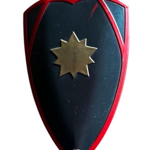 Pode incluir: Um escudo preto com borda vermelha e uma estrela dourada no centro.