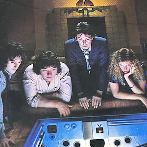May include: Four people, two men and two women, are looking down at a blue and silver control panel with buttons and a screen. The panel is on a wooden floor. The people are in a dimly lit room with a fireplace in the background.