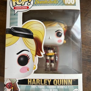 May include: A Harley Quinn vinyl figure from the DC Comics Bombshells series, number 166. The figure is in a clear display box. The box features the Pop! Heroes logo and the Harley Quinn name. The figure is wearing a costume with red, yellow, and brown colors.