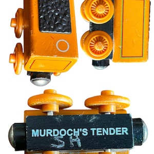Puede incluir: Un vagón de tender de tren de madera con los lados naranjas y ruedas negras. El tender tiene un vagón de carbón negro en la parte superior y un círculo blanco en el lateral. El tender tiene las palabras "Murdoch's Tender" impresas en la parte inferior.