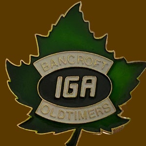 May include: A green maple leaf pin with a white oval containing the text "IGA" in black letters. The oval is surrounded by a white banner with the text "Bancroft Oldtimers" in gold letters.