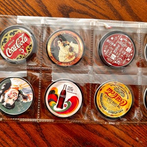 May include: Nine round Coca-Cola themed metal tokens in a clear plastic page protector. The tokens feature various Coca-Cola logos and imagery, including a woman drinking a Coca-Cola, a bottle of Coca-Cola, and the text "Drink Coca-Cola".