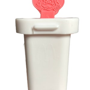 Könnte beinhalten: Weißer Plastik-Eis am Stiel-Formen mit einem rosa Charakterkopf oben drauf. Der Charakter hat ein rosa Gesicht mit schwarzen Augen und einem schwarzen Schnabel. Der Charakter hat schwarze Haare und trägt ein rosa Kleid.