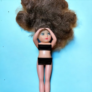 May include: A doll with brown hair and blue eyes is wearing a black bikini top and bottom. The doll is lying on its back with its arms raised above its head.