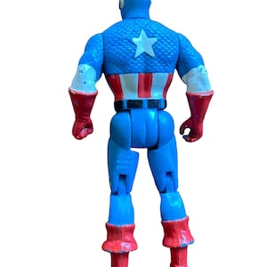 Puede incluir: Una figura de acci&oacute;n vintage de Capit&aacute;n Am&eacute;rica con un traje azul y rojo, una estrella blanca en el pecho y un cintur&oacute;n negro. La figura est&aacute; hecha de pl&aacute;stico y tiene brazos y piernas articulados. La figura est&aacute; de pie sobre un fondo blanco.