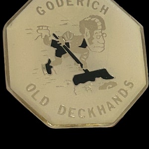 May include: A gold-colored, octagonal pin with a black and white illustration of a person holding a shovel. The text "GODERICH OLD DECKHANDS" is inscribed around the edge.