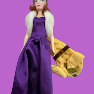 May include: A purple satin dress with a white fur collar and a yellow plush accessory. The doll has brown hair and blue eyes.
