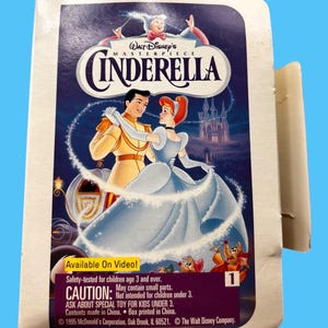 Puede incluir: Una caja de cartón blanca con una imagen de Cenicienta y el Príncipe Azul bailando. La caja es para un video de Walt Disney's Masterpiece Cenicienta. El texto "Available On Video!" está impreso en la caja. El texto "Safety-tested for children age 3 and over. May contain small parts. CAUTION: Not intended for children under 3. ASK ABOUT SPECIAL TOY FOR KIDS UNDER 3. Contents made in China. Box printed in China. © 1995 McDonald's Corporation, Oak Brook, IL 60521. © The Walt Disney Company." está impreso en la caja.
