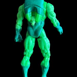 May include: A green action figure, seen from the back, with a backpack and helmet. The figure has a muscular physique and is posed with arms slightly extended. The legs are also visible, with a darker green shade at the feet.