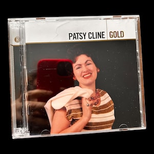 Op de afbeelding: Een cd-hoesje met het album "Gold" van Patsy Cline. De hoes toont een lachende vrouw in een gestreepte top en sieraden. De cd-hoes is van transparant plastic, met de albumtitel in gouden letters.