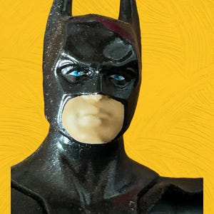 Puede incluir: Una figura de acción de plástico negro de Batman con una máscara azul y ojos azules. La figura lleva una capa negra y tiene una expresión seria.