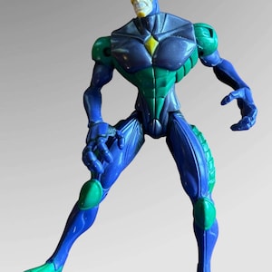May include: A blue and green action figure with a yellow and white mask. The figure is in a fighting pose with its arms raised.