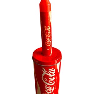 以下が含まれることがあります： 赤いコカ・コーラの缶の上に赤いろうそくが置かれています。ろうそくにはコカ・コーラのロゴが印刷されています。
