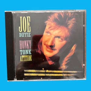 May include: A CD case with a black spine and a clear plastic front. The front features a photo of Joe Diffie, a country music singer, with long blonde hair and a mustache. The text on the case reads "Joe Diffie Honky Tonk Attitude".
