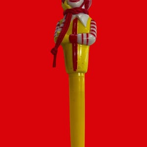 Puede incluir: Un bolígrafo de plástico amarillo y rojo con una figura de payaso en la parte superior. El payaso lleva una camisa a rayas rojas y blancas, un sombrero rojo y una bufanda roja.