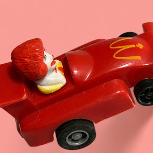 Puede incluir: Un coche de carreras de juguete rojo con un logotipo de McDonald's amarillo y una figura de payaso en el asiento del conductor.