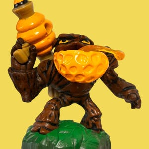May include: A brown action figure of a tree-like creature holding a honeycomb-shaped bowl and a honey dipper. The figure has a yellow bowl and a honey dipper. The figure is standing on a green base with a blue border. The background is yellow.