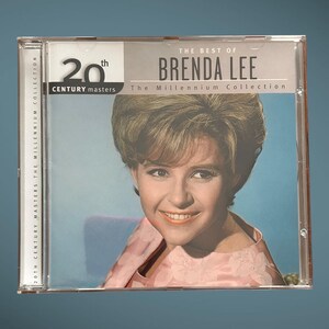 Peut inclure: Un boîtier de CD avec une photo de Brenda Lee, une chanteuse, sur la face avant. Le titre du CD est "The Best of Brenda Lee: The Millennium Collection". Le CD fait partie de la série 20th Century Masters.