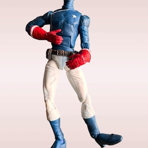 Pode incluir: Uma figura de a&ccedil;&atilde;o azul e branca de um super-her&oacute;i usando uma m&aacute;scara e luvas vermelhas e brancas. A figura est&aacute; em pose com um bra&ccedil;o levantado e o outro bra&ccedil;o dobrado no cotovelo. A figura est&aacute; em p&eacute; sobre um fundo branco.