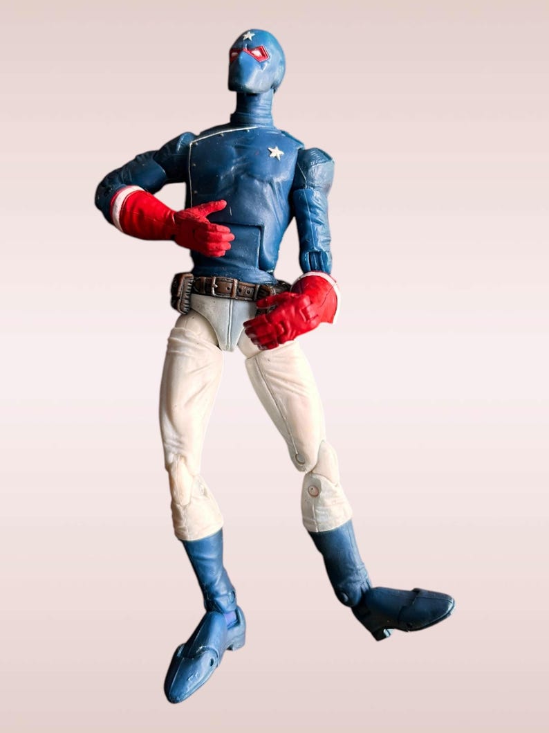 Pode incluir: Uma figura de a&ccedil;&atilde;o azul e branca de um super-her&oacute;i usando uma m&aacute;scara e luvas vermelhas e brancas. A figura est&aacute; em pose com um bra&ccedil;o levantado e o outro bra&ccedil;o dobrado no cotovelo. A figura est&aacute; em p&eacute; sobre um fundo branco.