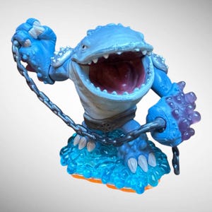 May include: A blue action figure of a creature with a wide open mouth, holding a chain. The figure has a light blue body, a white belly, and a dark red mouth. It stands on a blue and orange base.