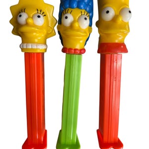 May include: Three vintage PEZ dispensers featuring The Simpsons characters: Lisa, Marge, and Bart. Each dispenser has a character head on top of a colorful, rectangular body. Lisa has a yellow head with a star-shaped hair, Marge has blue hair, and Bart has spiky yellow hair.