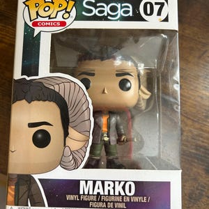 May include: A Pop! Comics Marko vinyl figure from the Saga series, displayed in its original packaging. The figure is of a character with tan skin, brown hair, and goat-like horns. The packaging includes the Pop! Comics logo and the name "Marko".