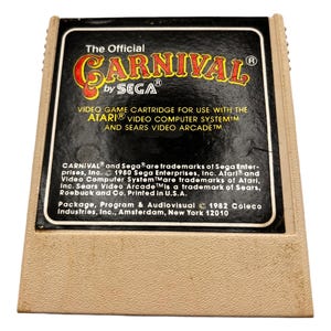 May include: A black Atari game cartridge with a beige label. The label reads "The Official CARNIVAL by SEGA" and "VIDEO GAME CARTRIDGE FOR USE WITH THE ATARI VIDEO COMPUTER SYSTEM AND SEARS VIDEO ARCADETM". The label also includes copyright information for Sega Enterprises, Inc., Atari, Inc., Sears, Roebuck and Co., and Coleco Industries, Inc.