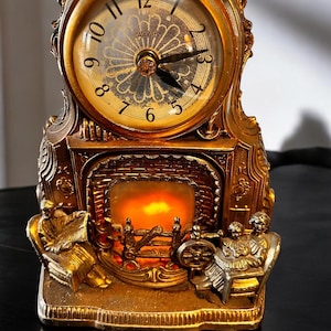 Op de afbeelding: Een goudkleurige mantelklok met een ronde wijzerplaat en Romeinse cijfers. De klok heeft een decoratief haardontwerp met een warm, gloeiend licht. Twee figuren zitten aan weerszijden van de haard.
